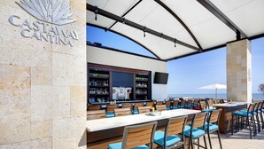 Zona de bar con pared de piedra, una pantalla grande y vista al océano.