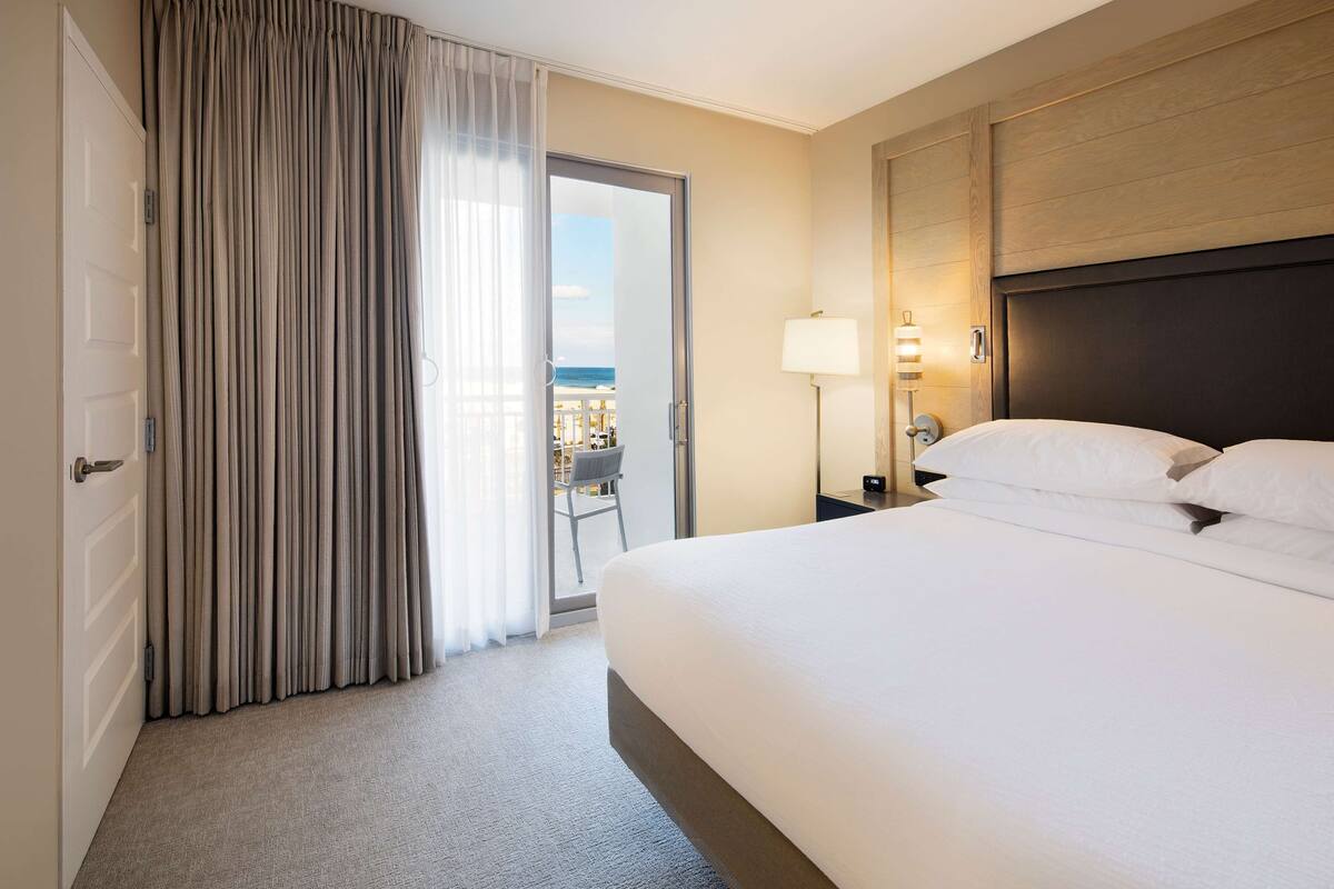 Une chambre d’hôtel avec un lit, une table de chevet, une lampe, une vue sur le balcon et une porte avec des rideaux.