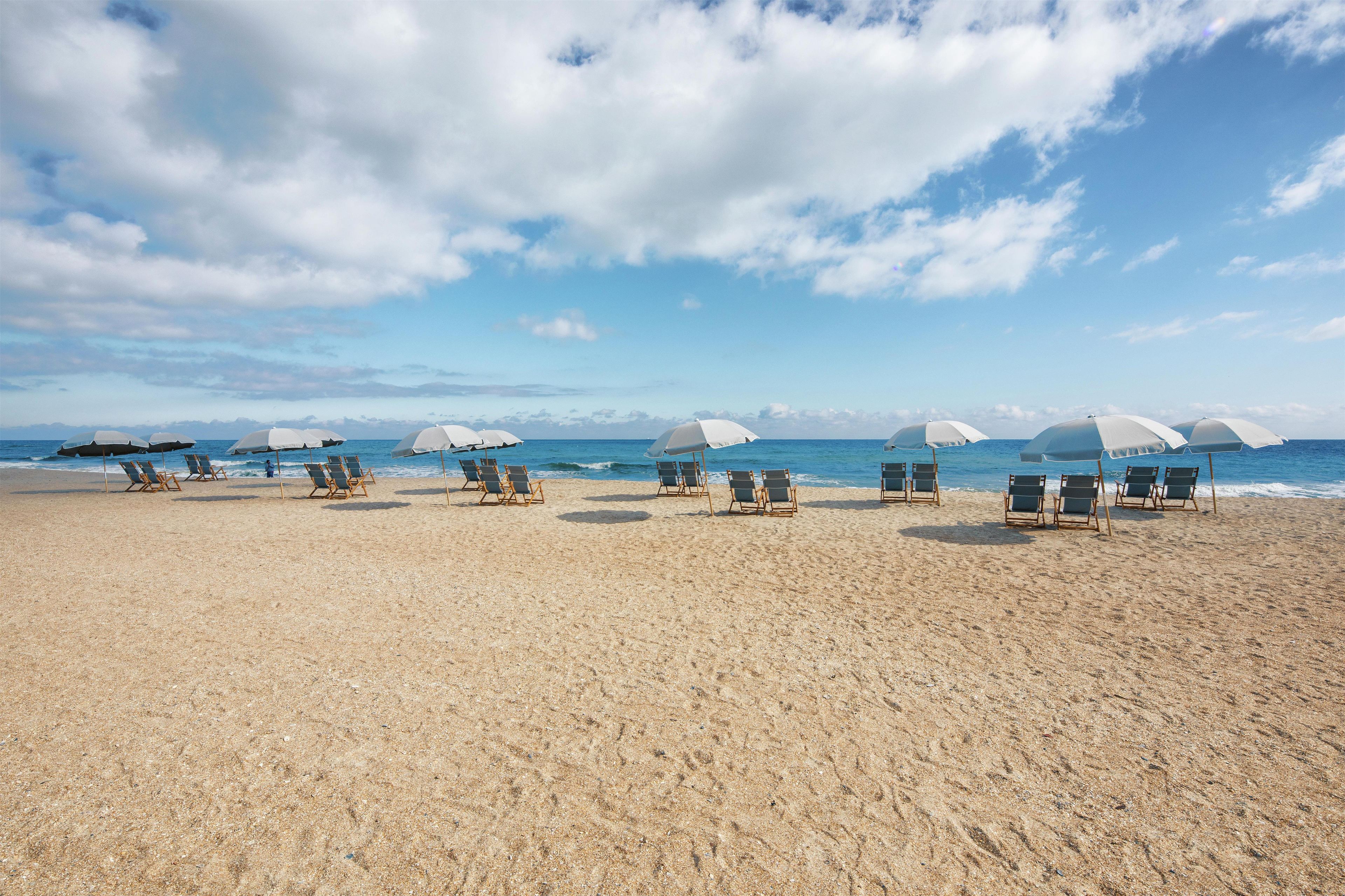 Ein Strand mit Reihen von Liegestühlen und Sonnenschirmen, einem klaren Himmel und dem Meer im Hintergrund.