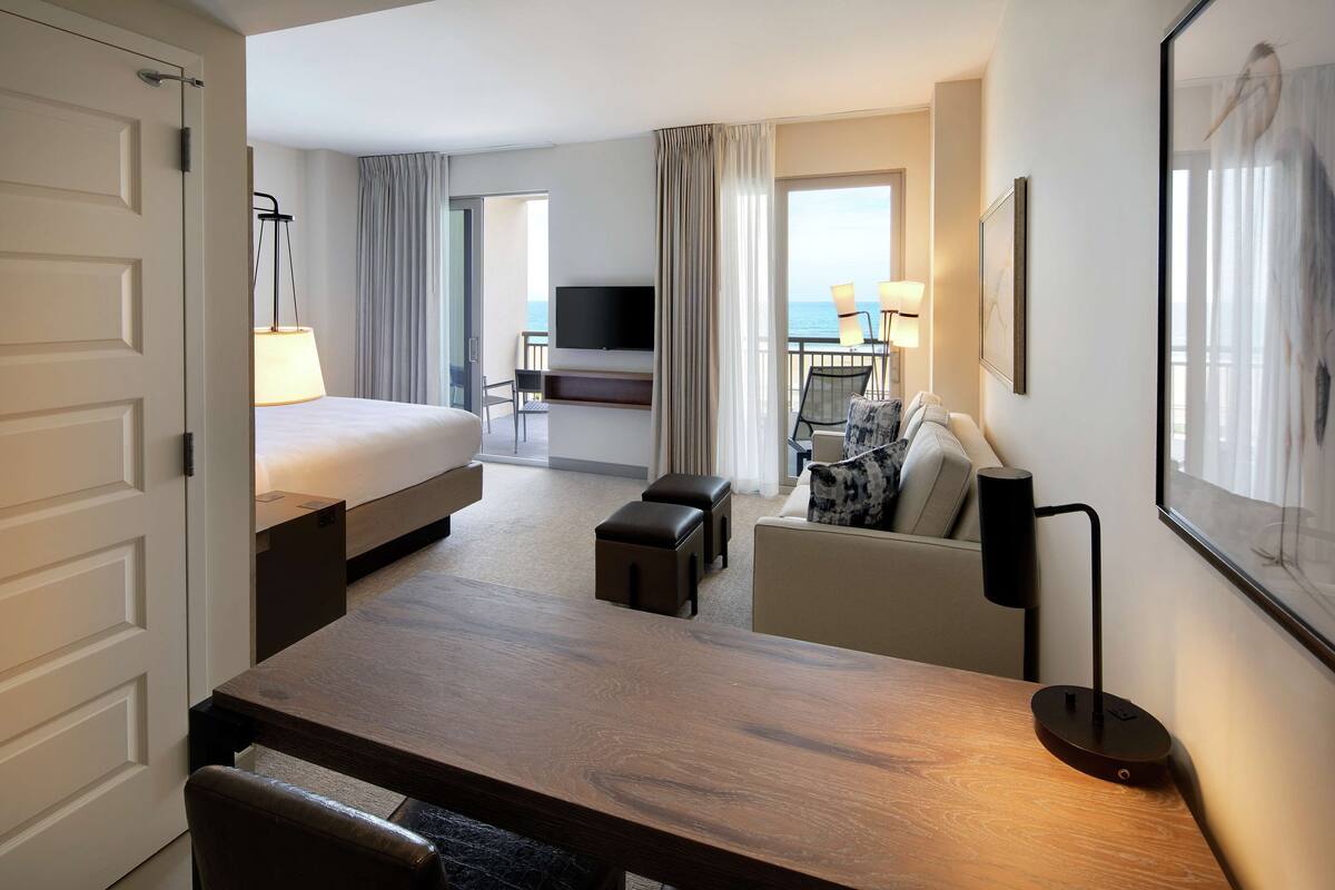 Une chambre d’hôtel avec un lit, un bureau, une chaise, une télévision et un balcon donnant sur la mer.