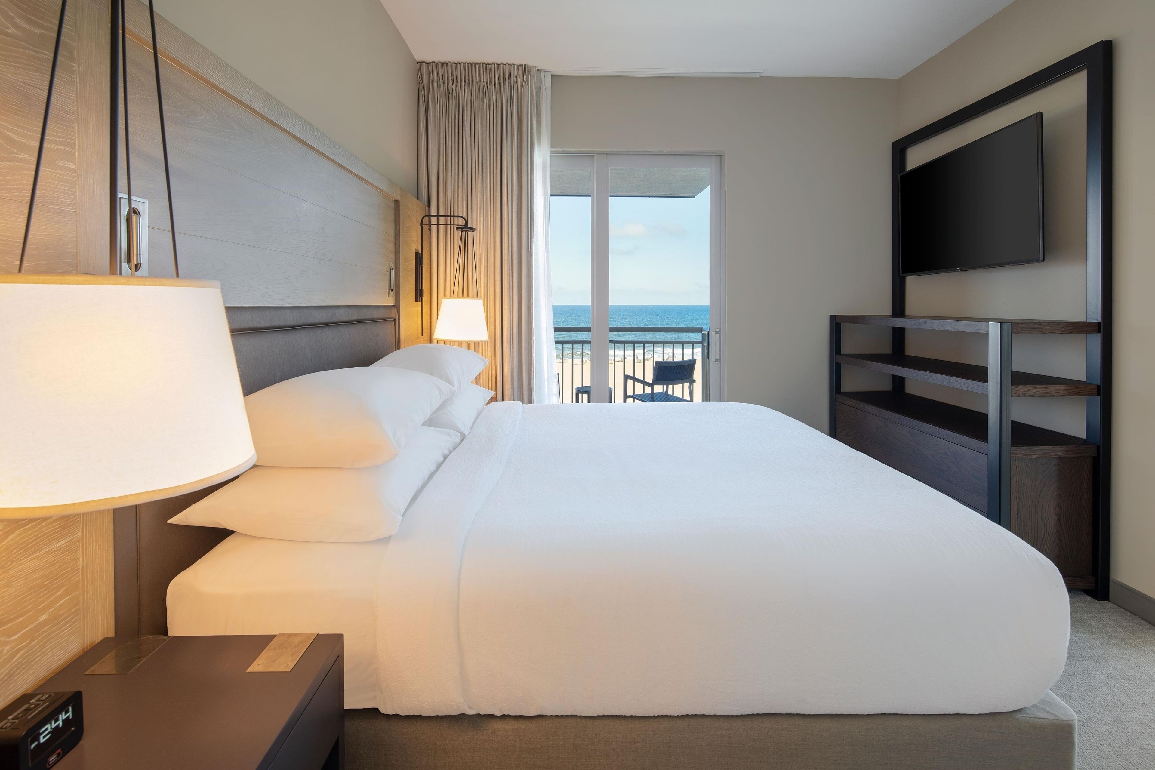 Ein Hotelzimmer mit einem großen Bett, einem Fernseher, einem Schreibtisch mit Lampe und Blick aufs Meer.