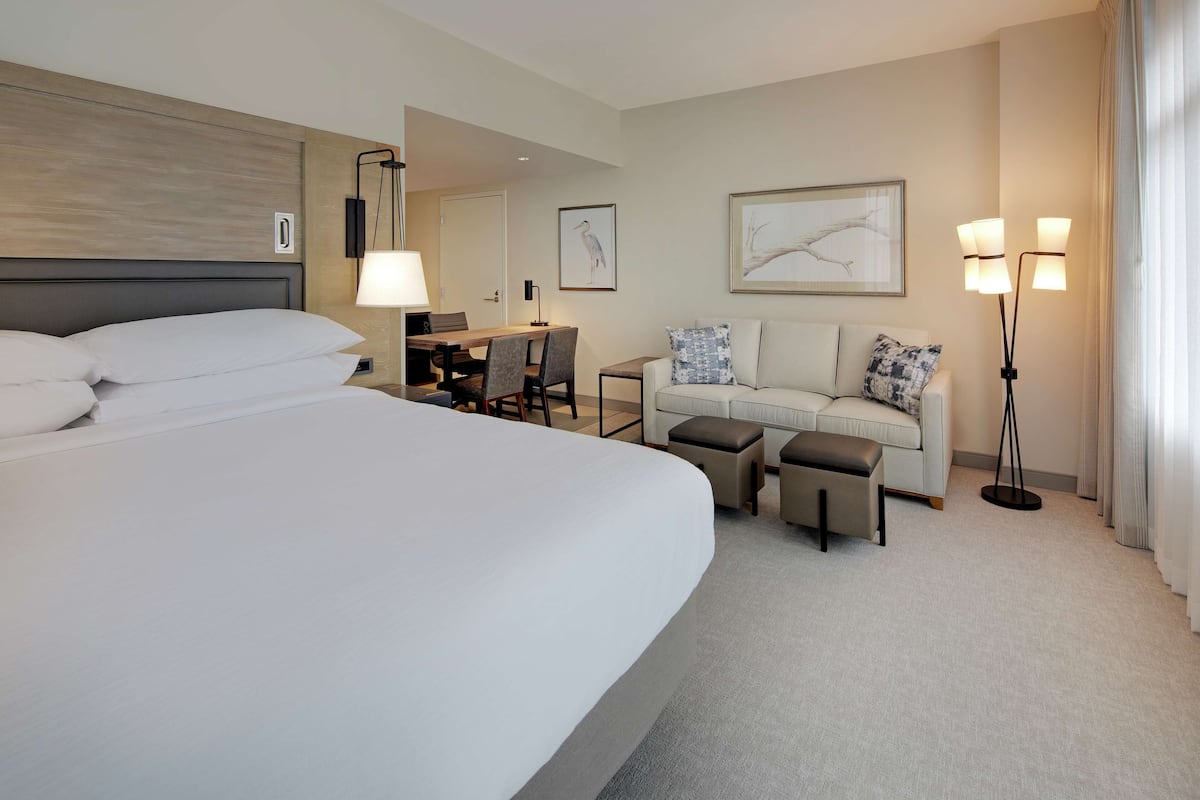 Une chambre d’hôtel avec un grand lit, un bureau avec une chaise, un canapé avec des coussins et deux lampadaires.