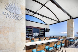 Angolo bar con vista sull'oceano, caratterizzato da una parete in pietra con la scritta 'CASTAWAY CANTINA' e un bancone fornito di diverse bottiglie.
