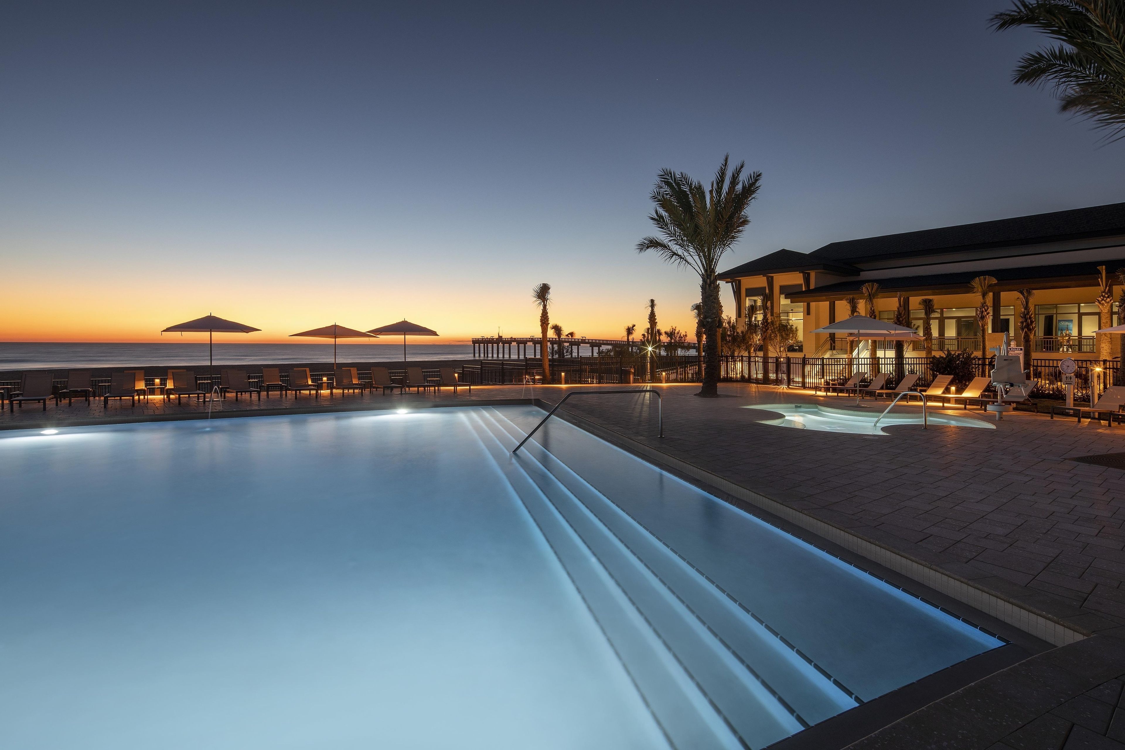 Une piscine avec des chaises longues et des parasols, un bâtiment doté d’un balcon, et une vue sur l’océan et les montagnes au crépuscule.