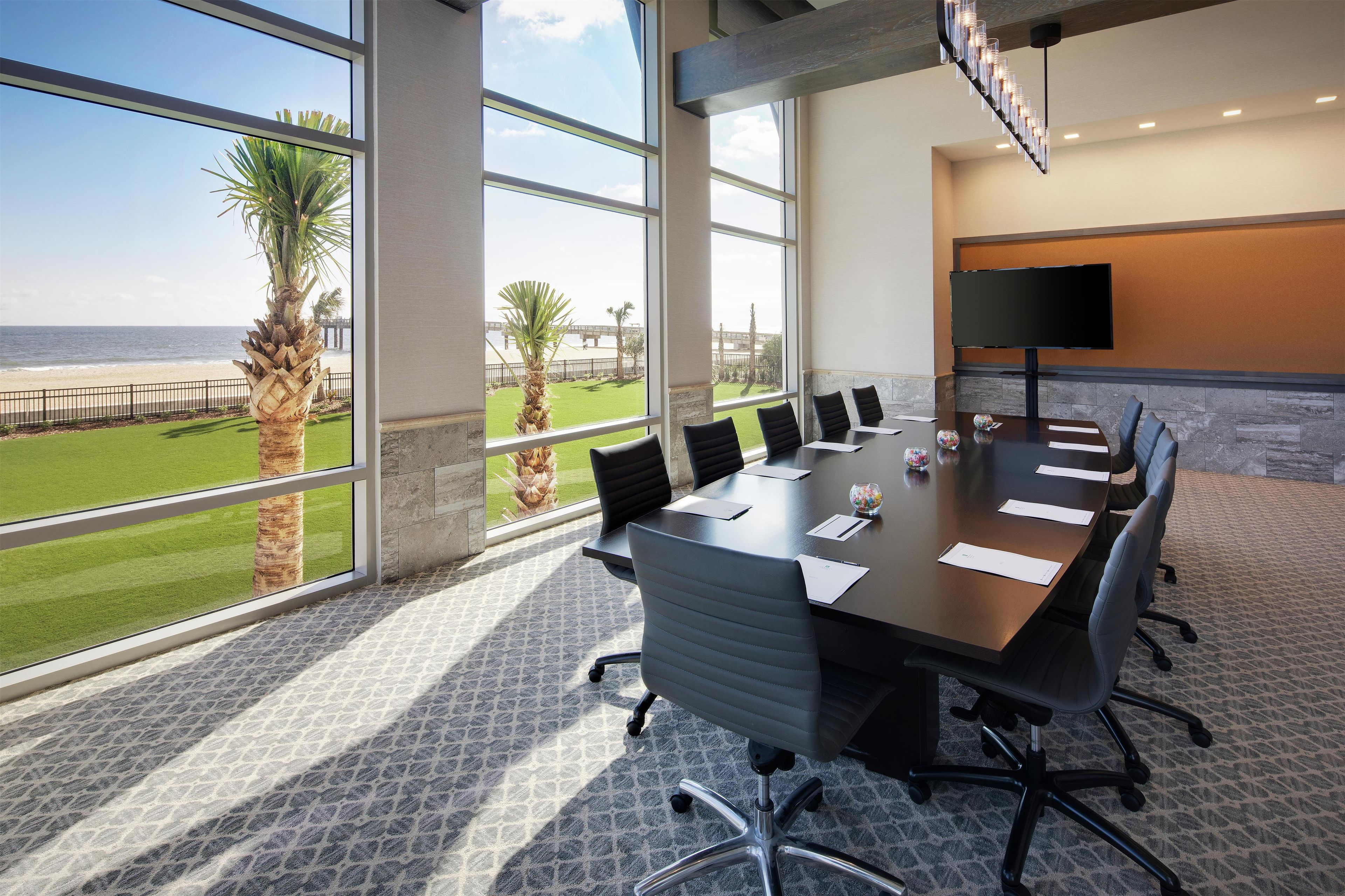 A conference room with a large table, chairs, a TV, and a view of the beach.