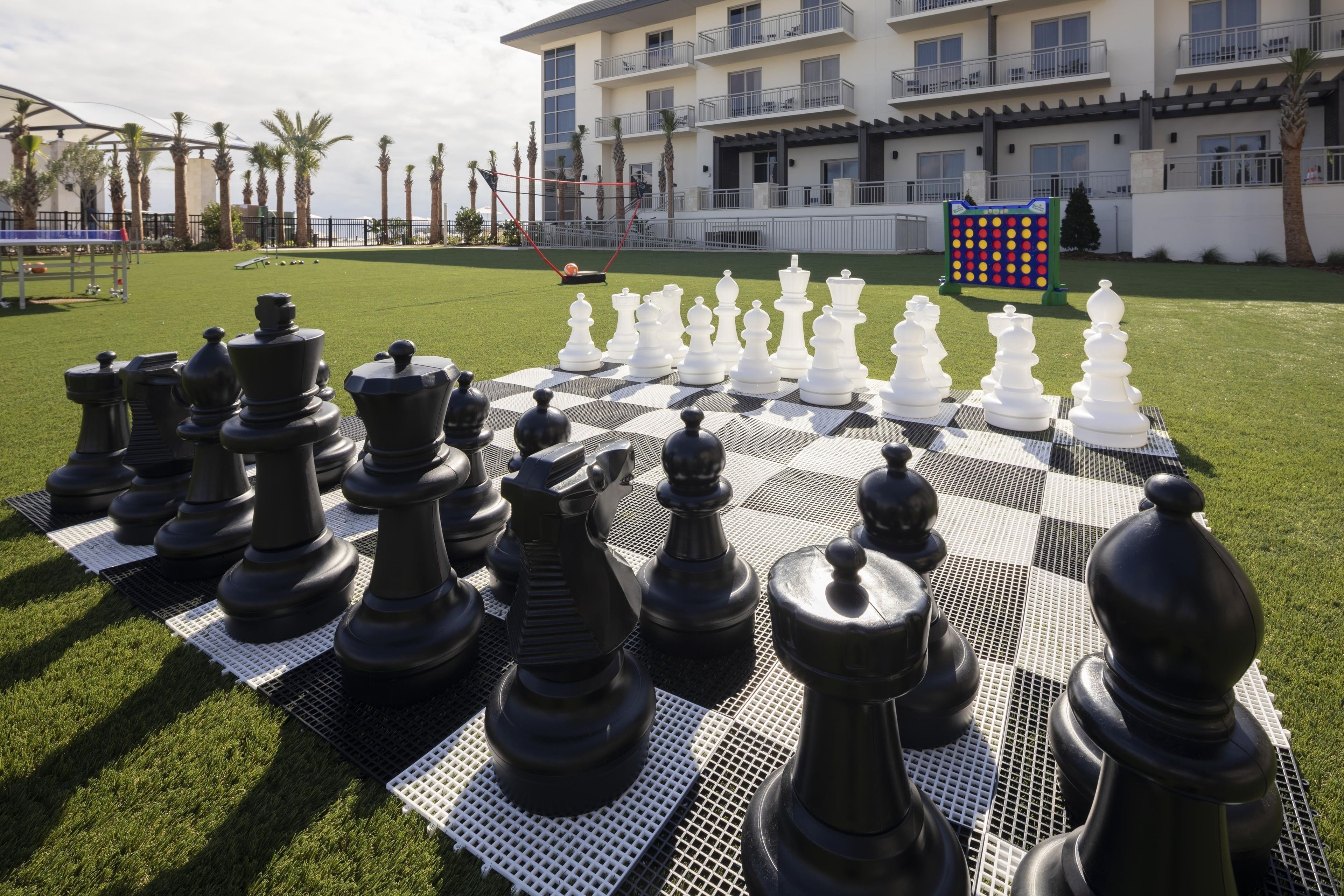A large outdoor chess set on a checkered table in a courtyard with a building in the background.