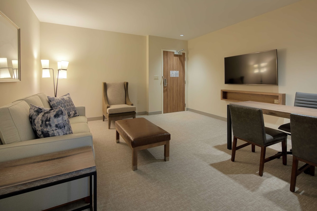 Une chambre d’hôtel comprenant un canapé, un banc, une table à manger avec des chaises, un téléviseur à écran plat fixé au mur et une porte avec un panneau en bois.