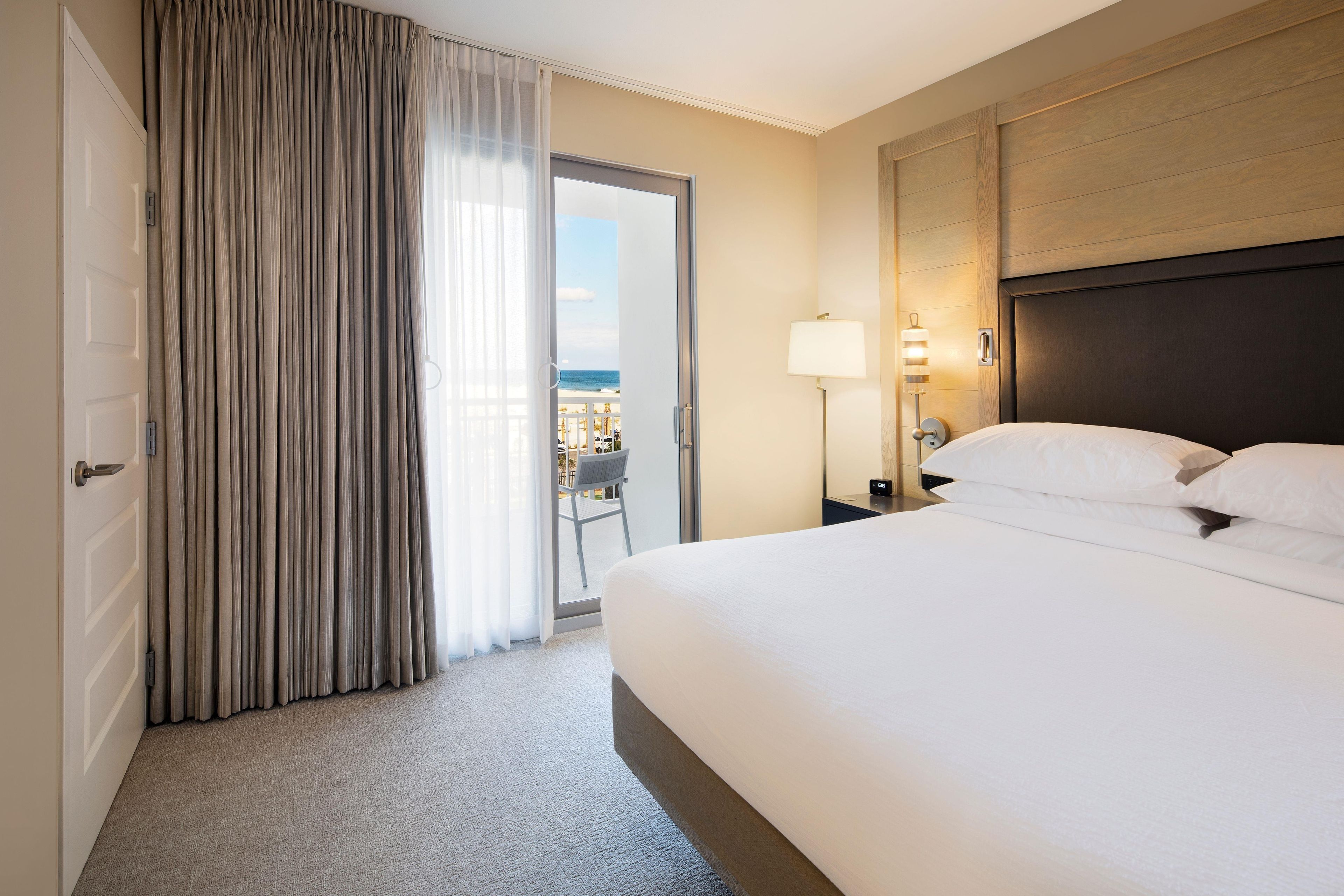 A hotel room with a bed, a nightstand with a lamp, a view of the balcony, and a door with curtains.
