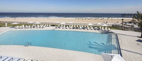 A pool area with lounge chairs and umbrellas, a beach with umbrellas and people, and a pier in the background.