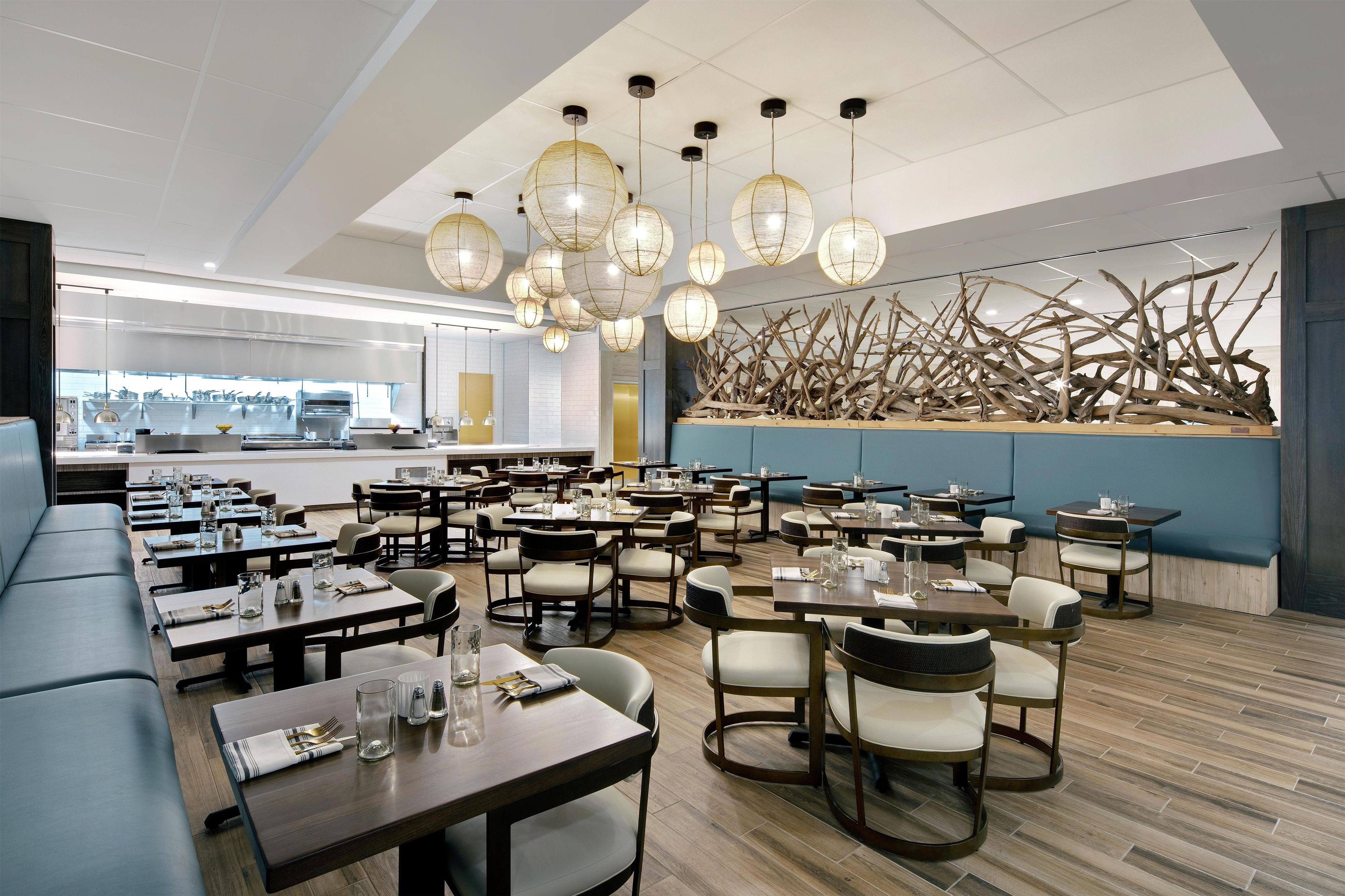 A modern restaurant with multiple dining tables and chairs, a kitchen area in the background, and a decorative wall with natural elements.