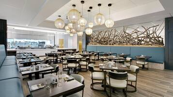 Un restaurante moderno con varias mesas y sillas, un área de cocina al fondo y una pared decorativa con elementos naturales.