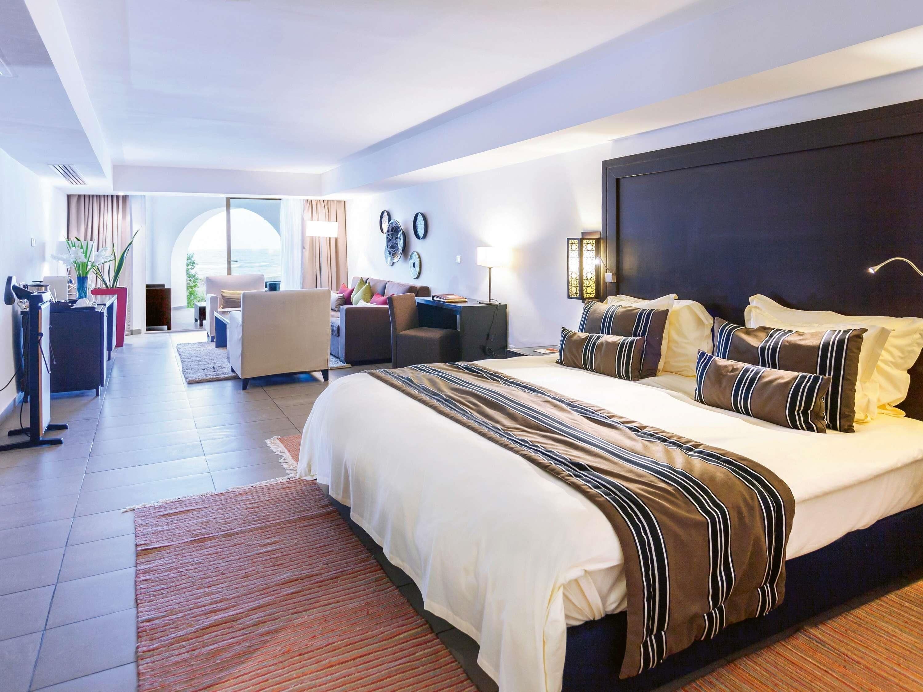 A modern hotel room with a large bed, a sitting area with a sofa and armchairs, a desk, and a view through a large window.