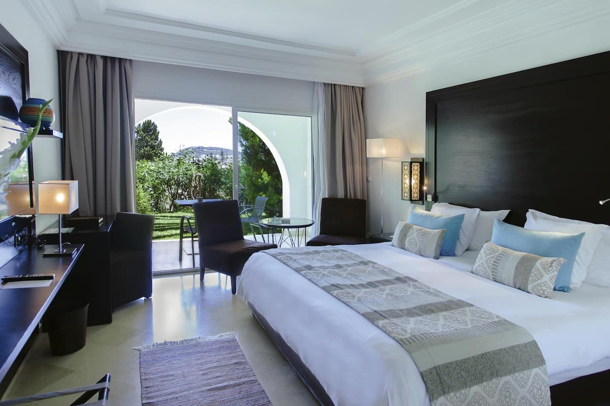 Une chambre d’hôtel moderne avec un grand lit, un bureau et une chaise. La fenêtre offre une vue sur la verdure environnante.