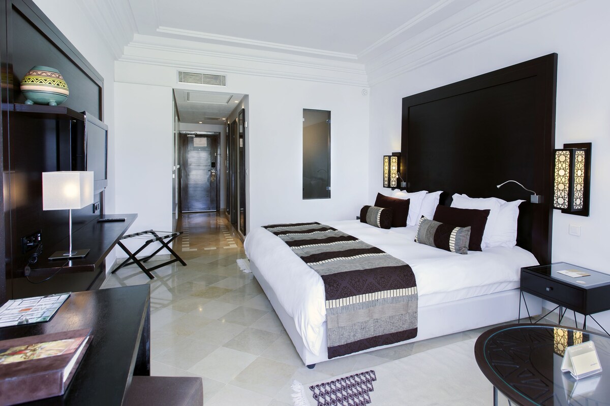A modern hotel room with a large bed, a desk, and a chair. There is a door leading to the hallway.