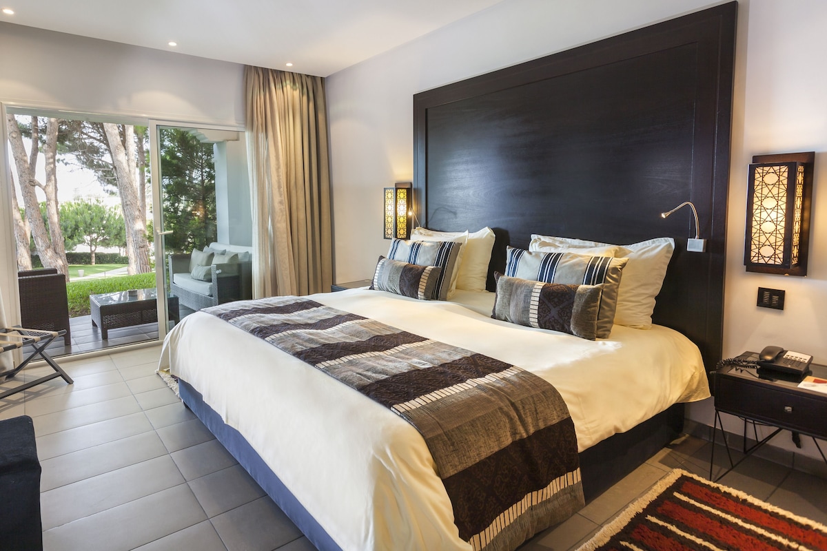 A modern hotel room with a large bed, a desk, and a balcony with a view of trees.