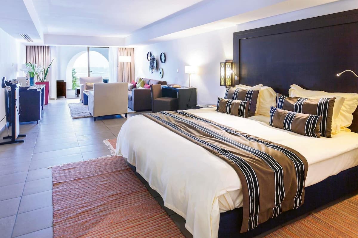 A modern hotel room with a large bed, a sitting area with a sofa and armchairs, a desk, and a view of the outdoors.