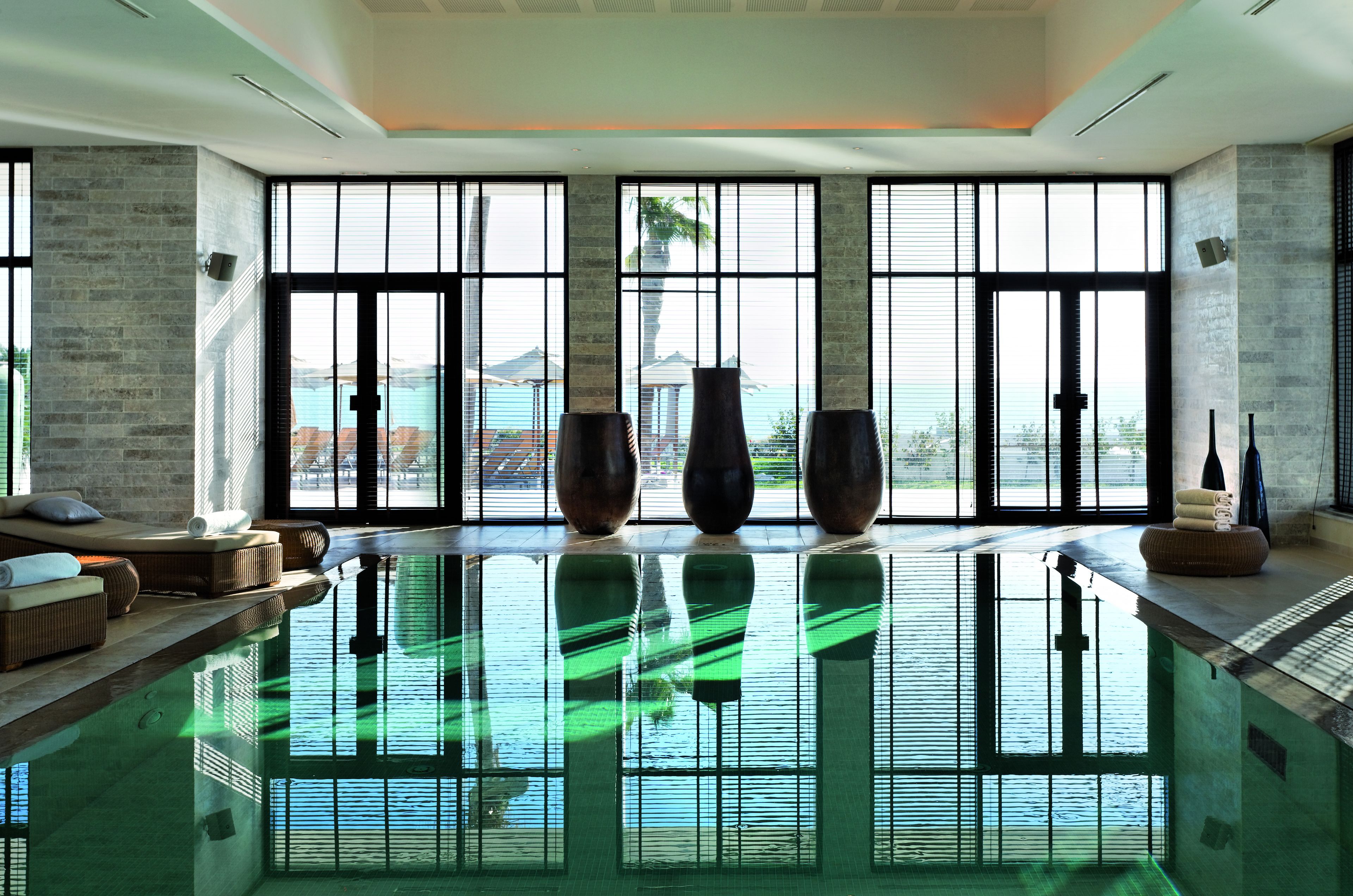 Una piscina interior moderna con ventanas amplias, un área de descanso con asientos y jarrones decorativos.