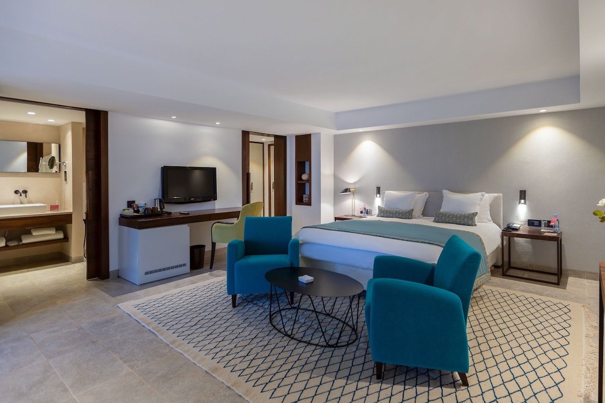 A modern hotel room with a bed, a desk with a TV, a bathroom, and a seating area with chairs.