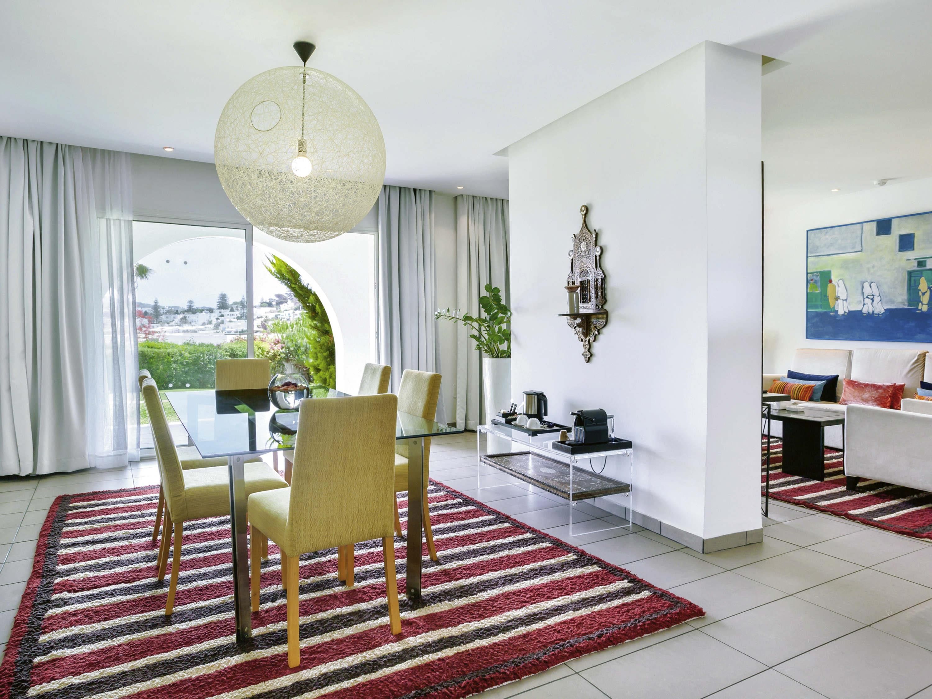 A modern living room with a dining area, a large window with curtains, a hanging light fixture, and a painting on the wall.