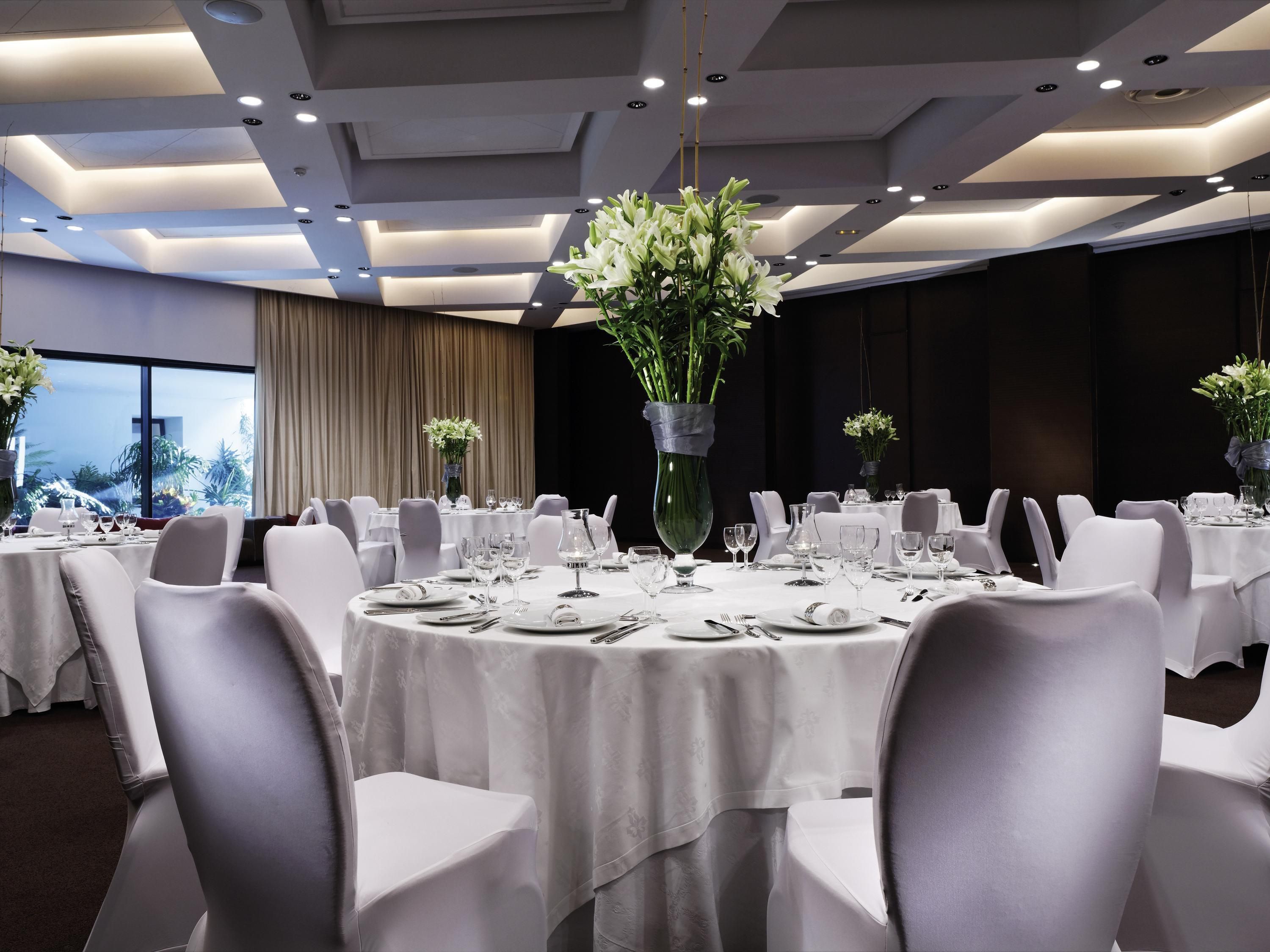 Un salón de banquetes con mesas redondas dispuestas para comer, sillas blancas y centros de mesa florales.