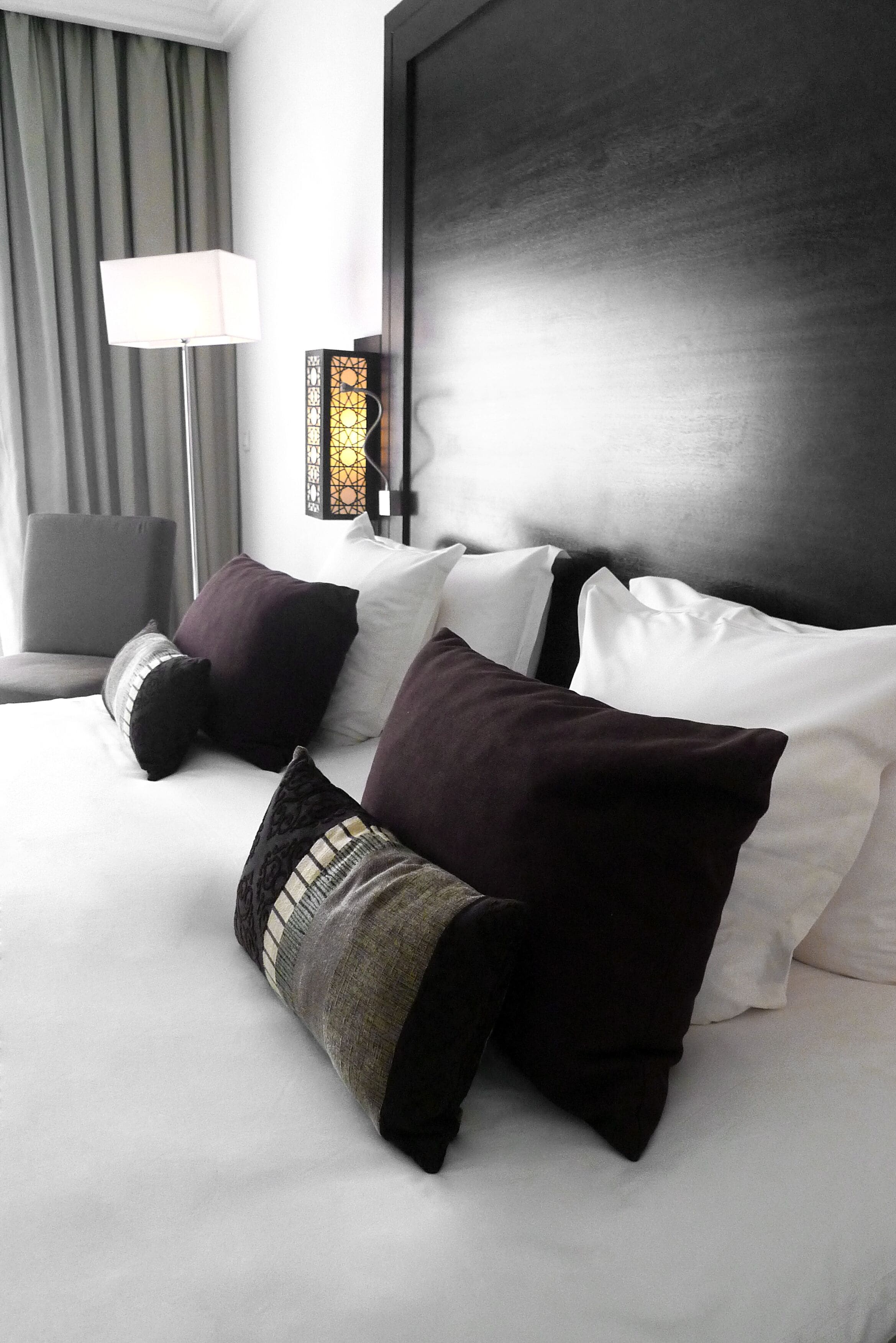 Una cama bien hecha con almohadas blancas y oscuras, un cabecero moderno y una lámpara de pie.