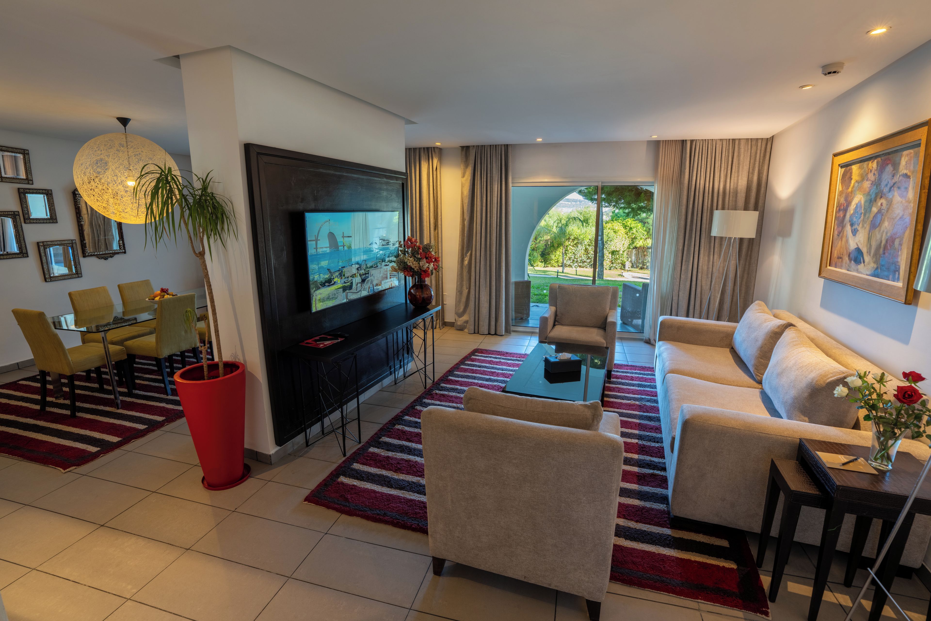 Uma sala de estar moderna com televisão de tela plana, assentos confortáveis, área de refeições e uma grande janela com vista para o verde.