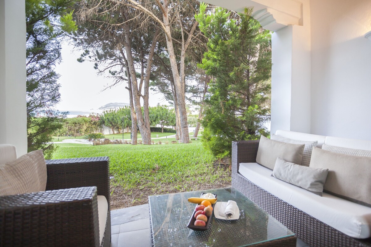 A modern outdoor seating area with wicker furniture, a glass table with fruits and a napkin, and a view of a lush garden and distant landscape.