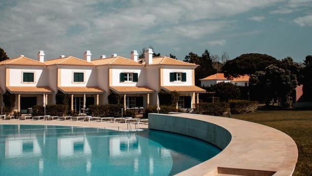 Uma piscina com borda curva, um edifício com telhado em azulejos e uma área gramada.