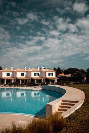 A swimming pool with a curved edge, a building with a tiled roof, and a grassy area. - Vila Bicuda Resort (Cascais)