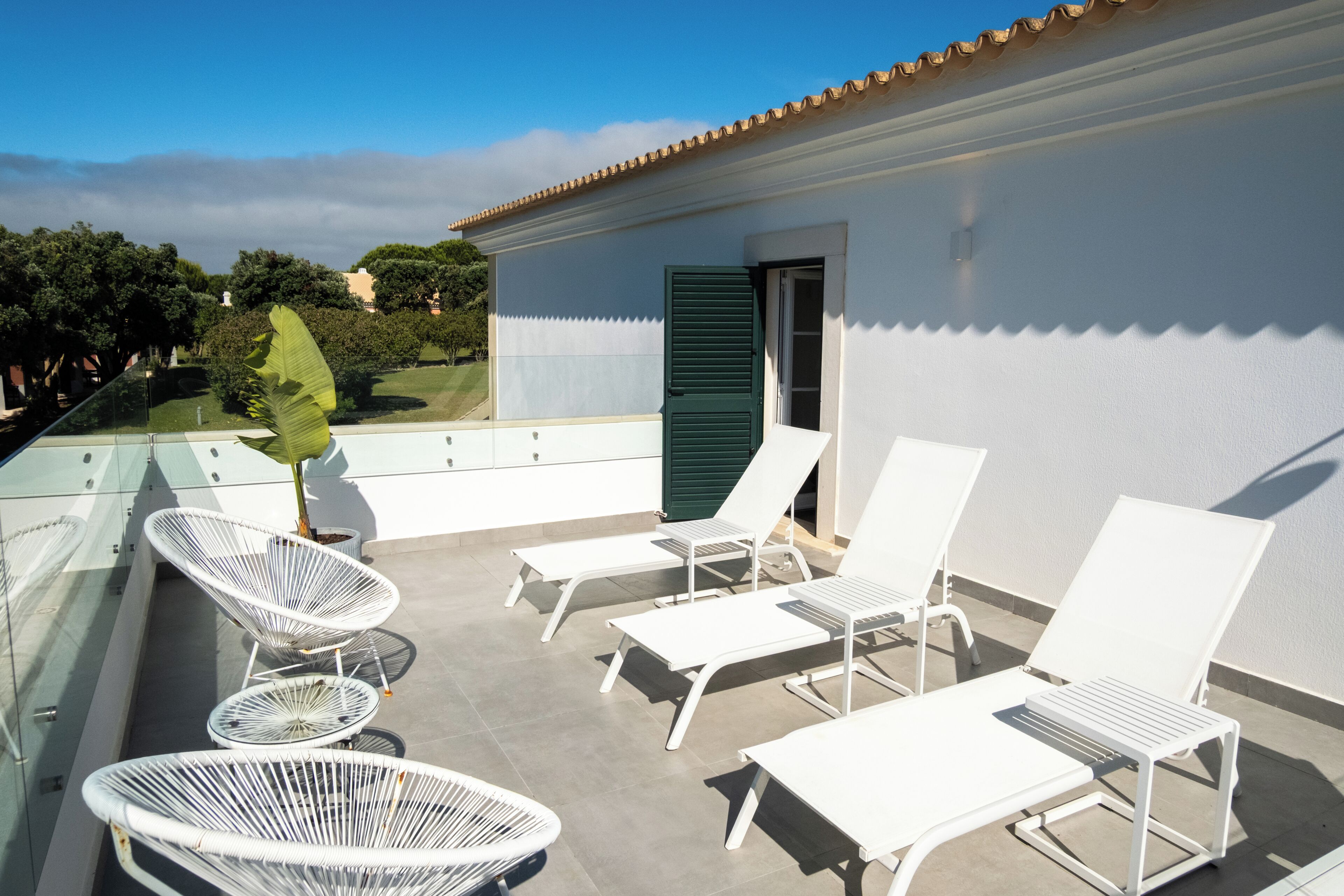 Un balcón con sillones blancos de descanso y una planta en maceta, con vista a una zona residencial.