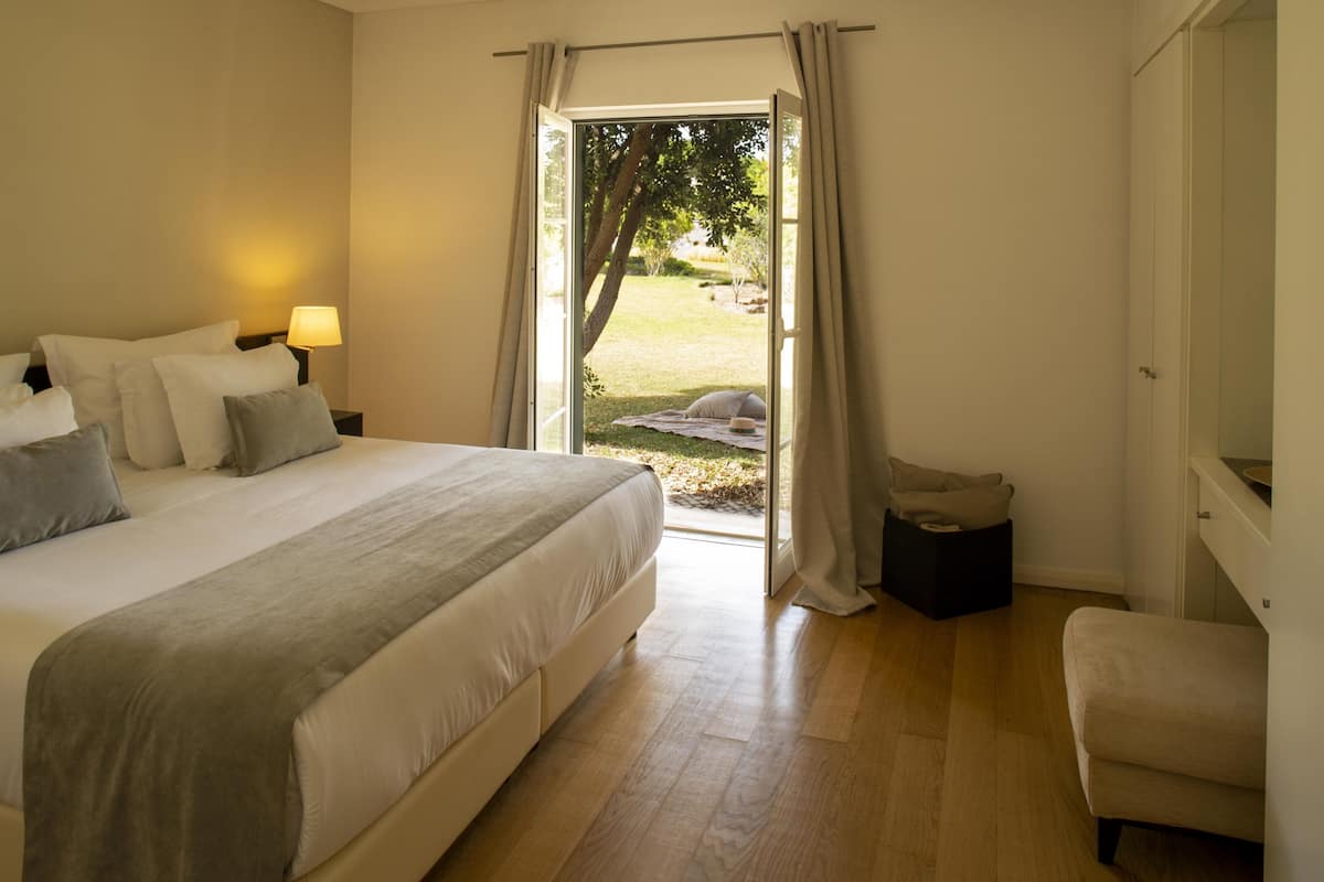 Une chambre avec un grand lit, une table de chevet avec une lampe, une chaise et une vue sur un espace extérieur, accessible par une porte ouverte.