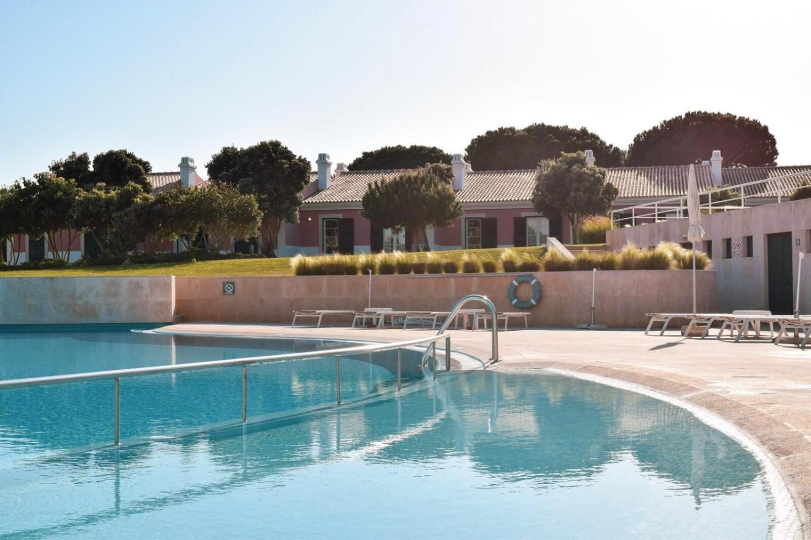 Une piscine à l’eau d’un bleu cristallin, entourée d’une terrasse en béton où sont disposées des chaises longues et où l’on peut apercevoir une bouée de sauvetage fixée au mur.