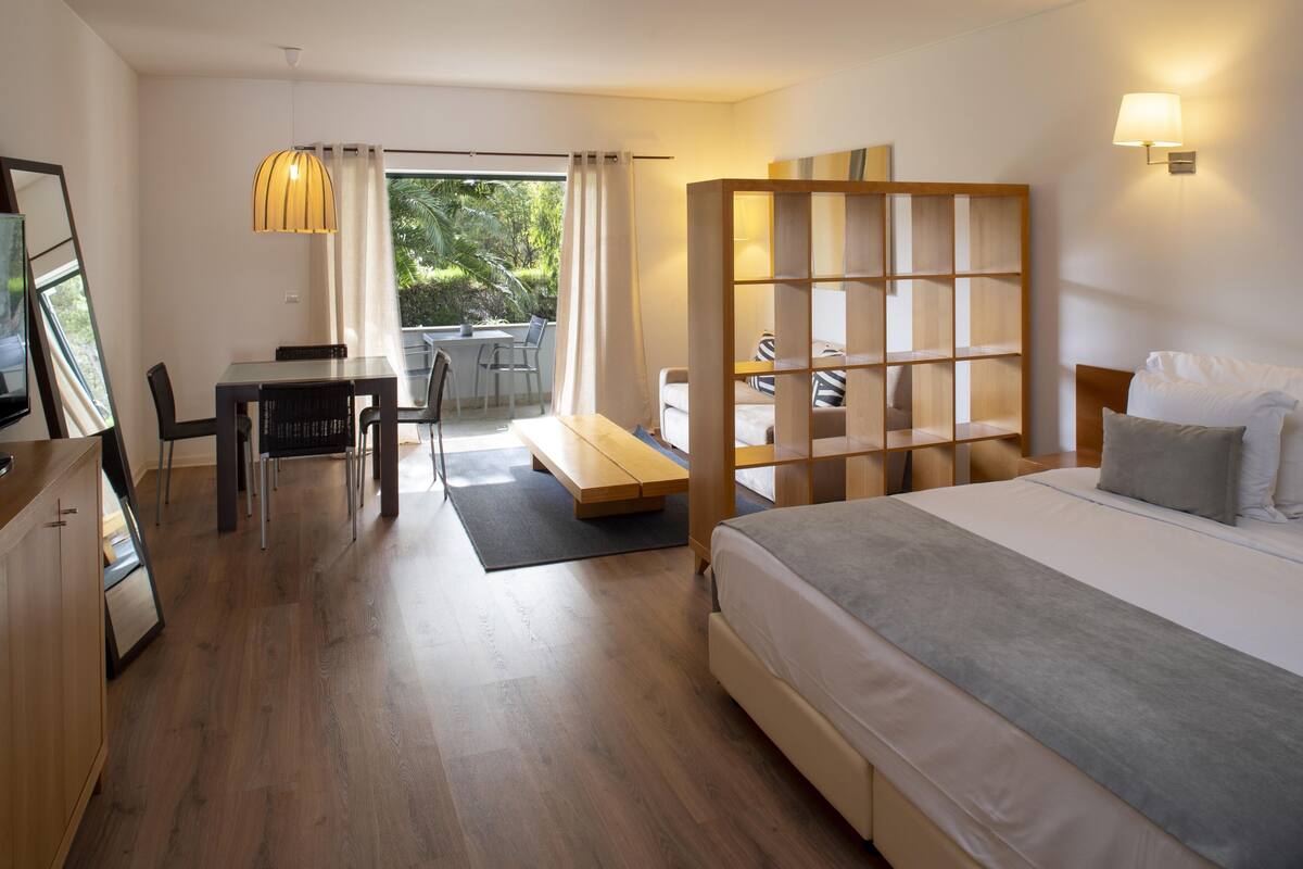 Une chambre d’hôtel comprenant un lit, un bureau, une chaise, un banc, une bibliothèque en bois et une vue sur l’extérieur.
