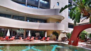 A pool area with lounge chairs and umbrellas, surrounded by a modern building with large windows. - Lancaster Tamar Hotel (Hadath)