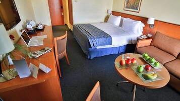 A hotel room with a bed, a desk with a laptop, a dining table with fruit, and a sofa.