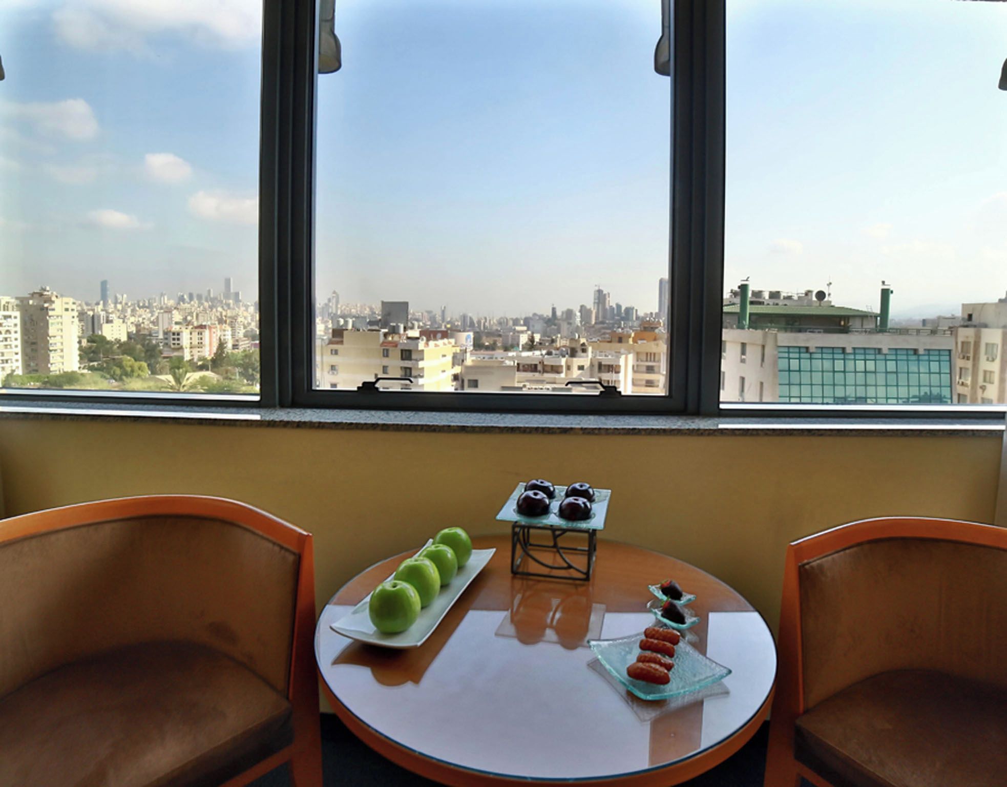 A room with a table and chairs, a window with a city view, and a plate with apples and pastries.
