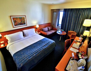 A hotel room with a large bed, a sofa, a desk with a laptop, a small table with fruits, and a window with curtains. - Lancaster Tamar Hotel (Hadath)