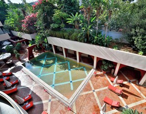 Une piscine sur le toit, dont le fond est décoré de motifs géométriques en mosaïque, entourée de chaises longues rouges et de plantes en pot.
