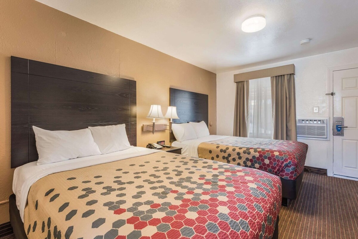 A hotel room with two beds, a nightstand with lamps, a window with curtains, and a door with an intercom.