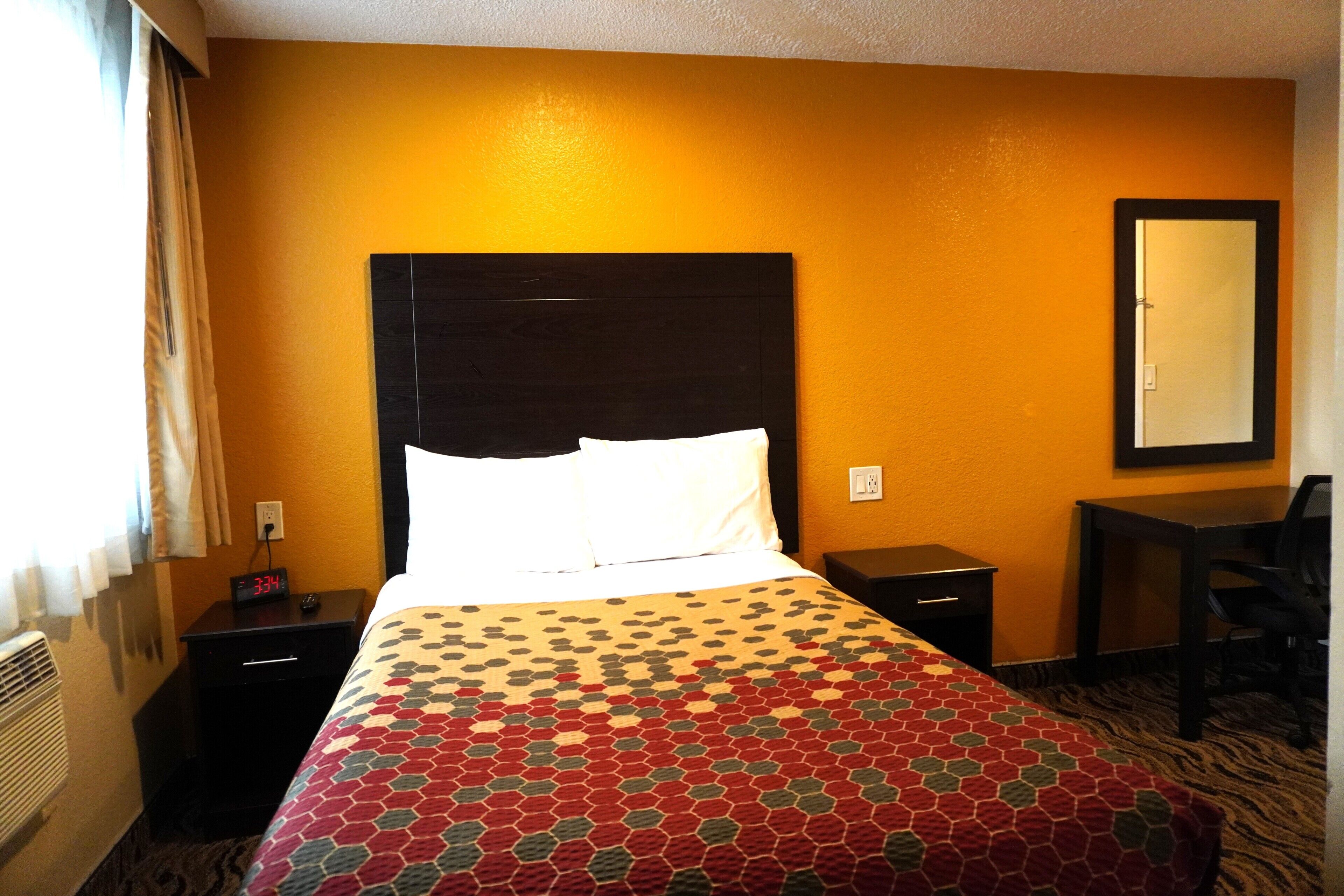 A hotel room with a bed, a nightstand with a clock, a desk, and a chair.
