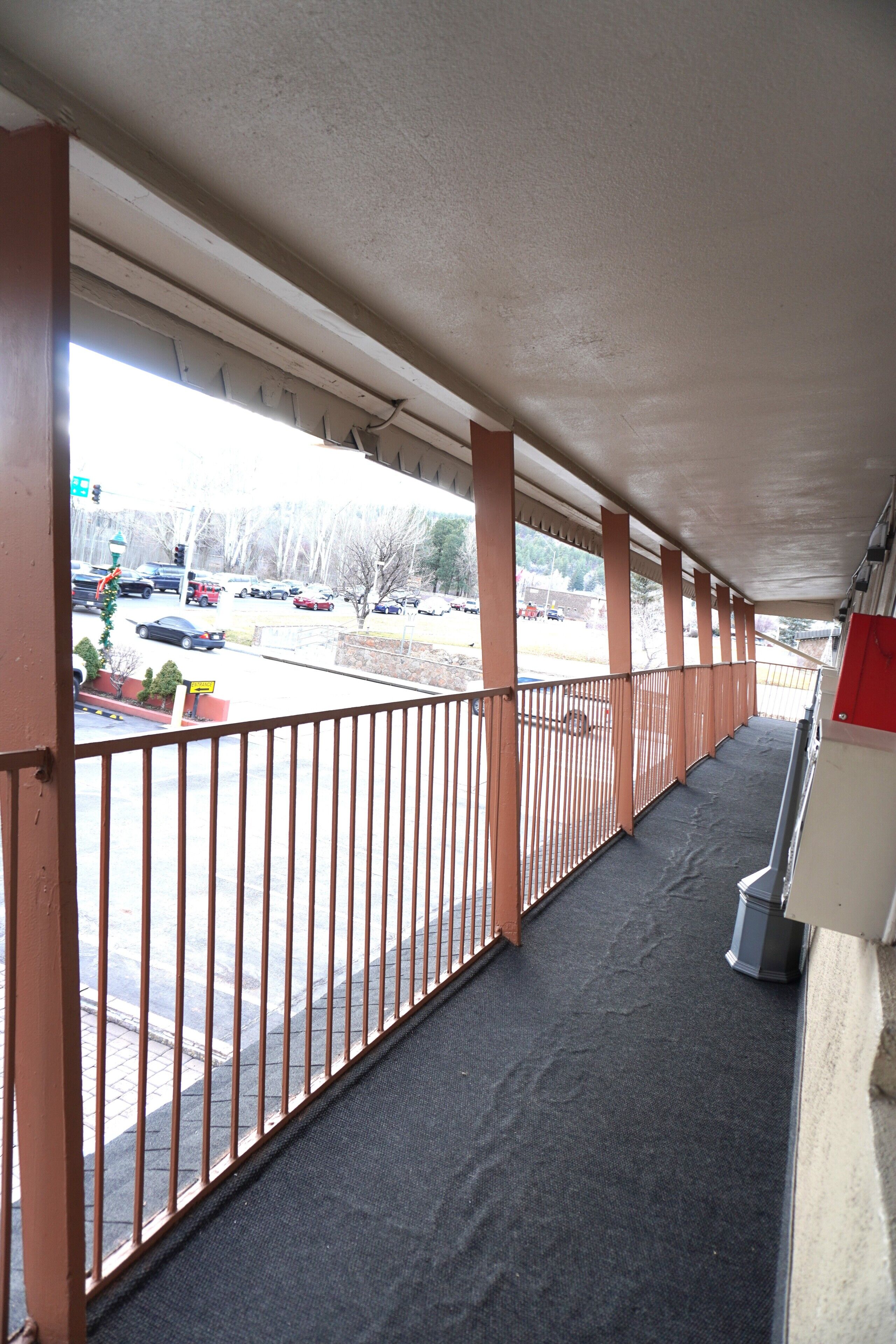 A covered outdoor area with a metal railing, a concrete floor, and a view of parked cars and trees.