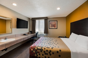 Quarto de hotel com cama, escrivaninha, cadeira, pia, espelho, TV de tela plana e janela com cortinas.