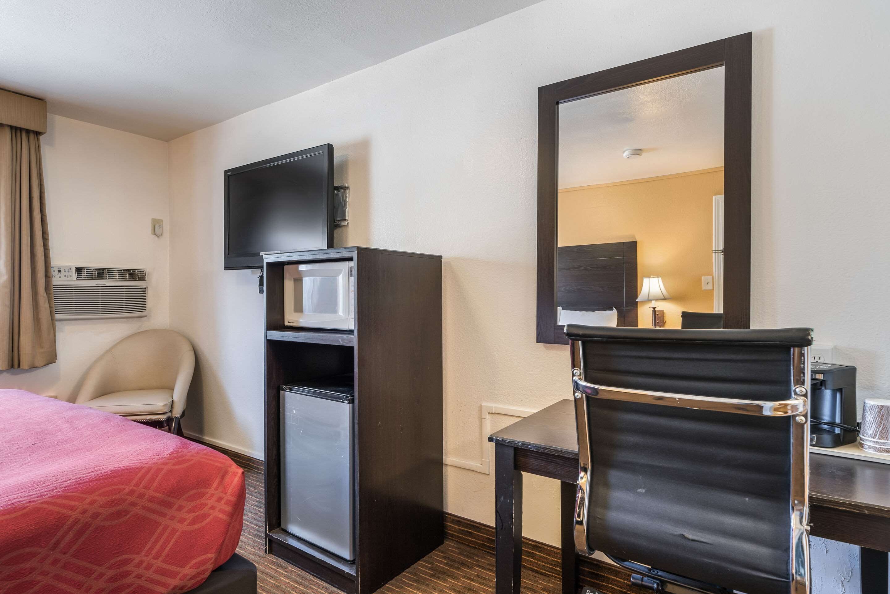 A hotel room with a bed, a desk with a chair, a television, a refrigerator, and a mirror.