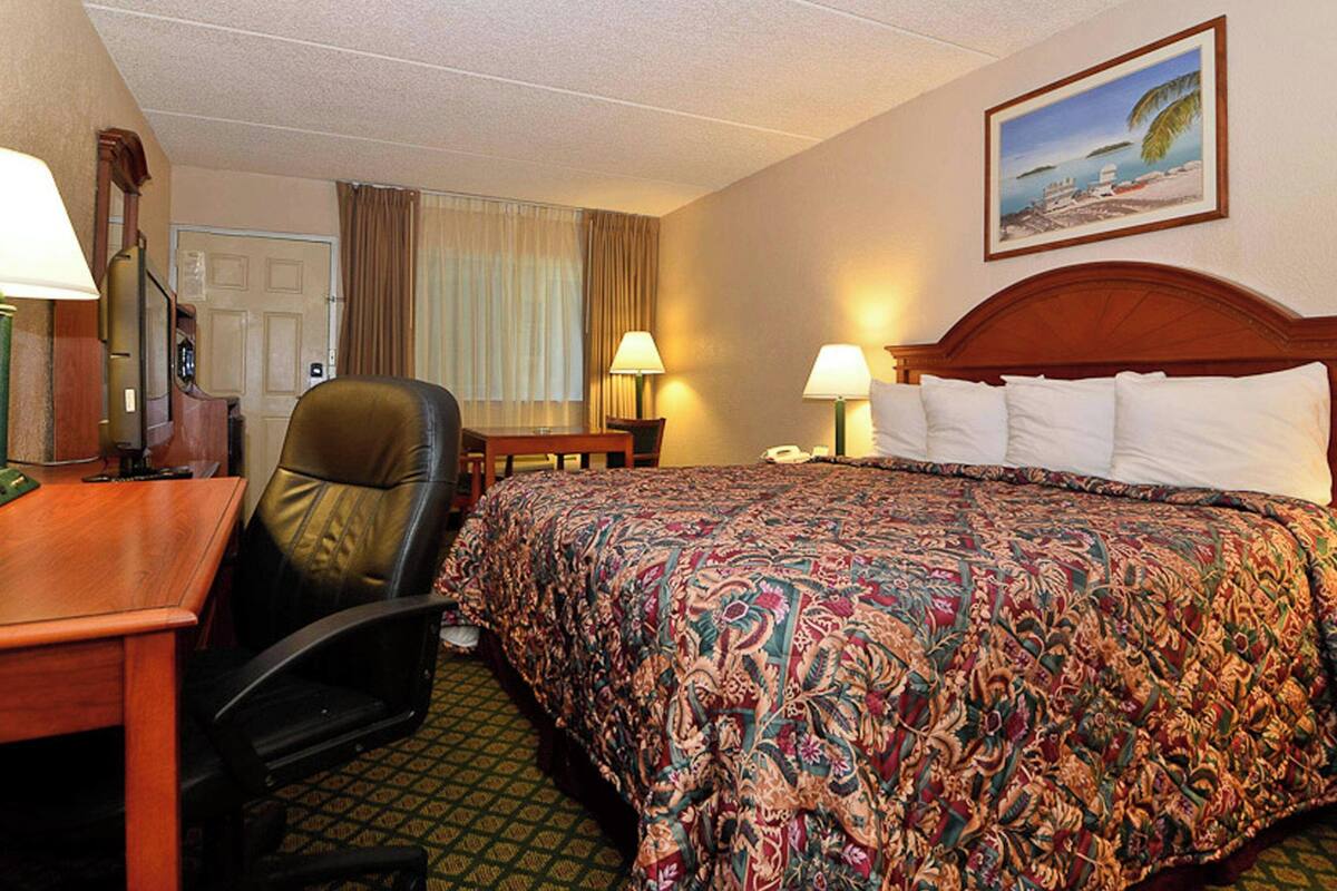 A hotel room with a bed, a desk, a chair, two lamps, a painting, and a door.