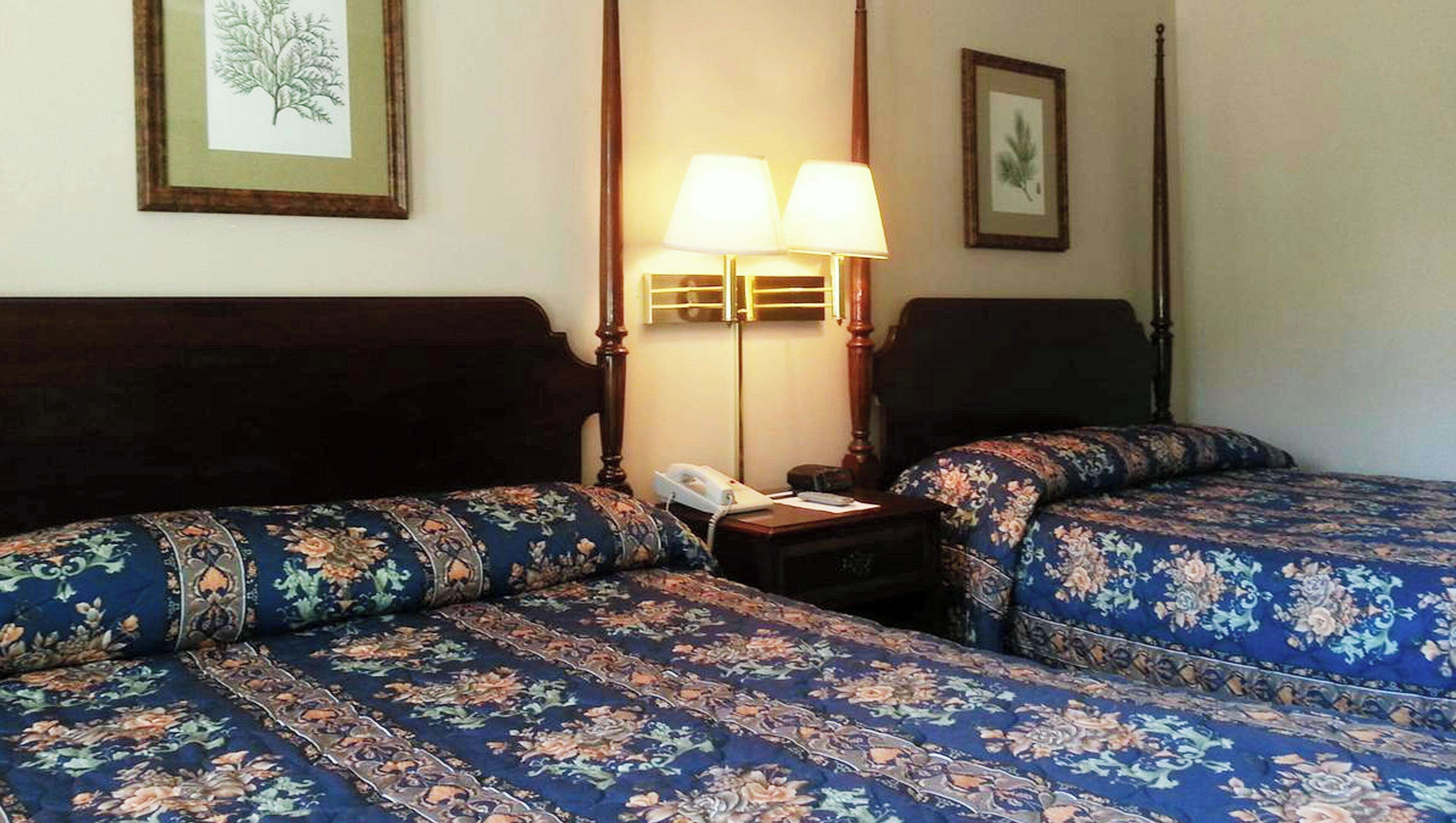 A hotel room with two beds, each with a floral-patterned bedspread, a nightstand with a lamp, and framed botanical artwork on the walls.