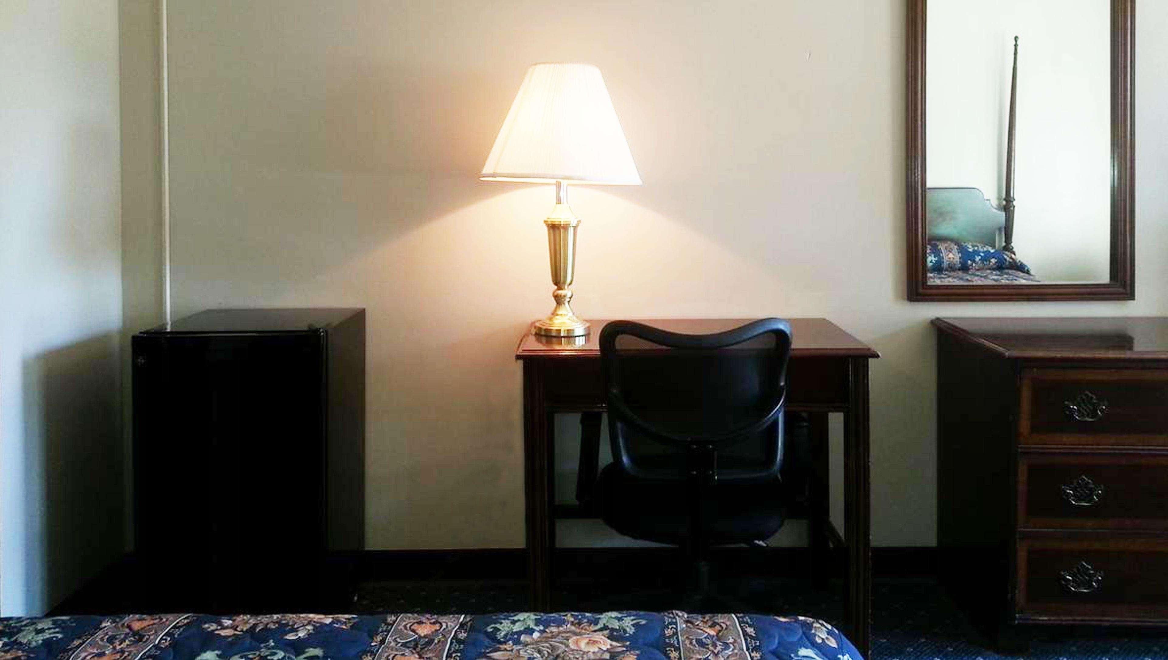 Habitación de hotel con escritorio, silla, lámpara, cómoda y cama con colcha estampada.