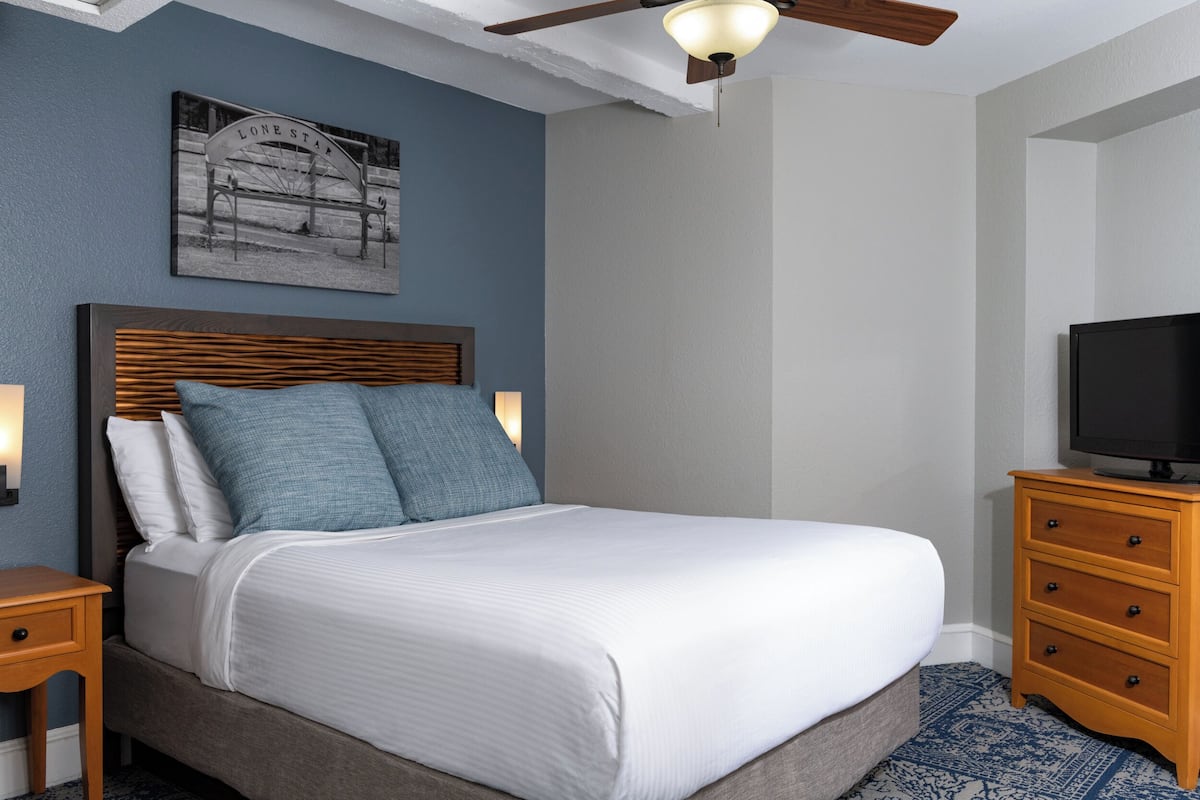 A neatly arranged bedroom with a bed, a wooden nightstand, a dresser, a television, and a ceiling fan.