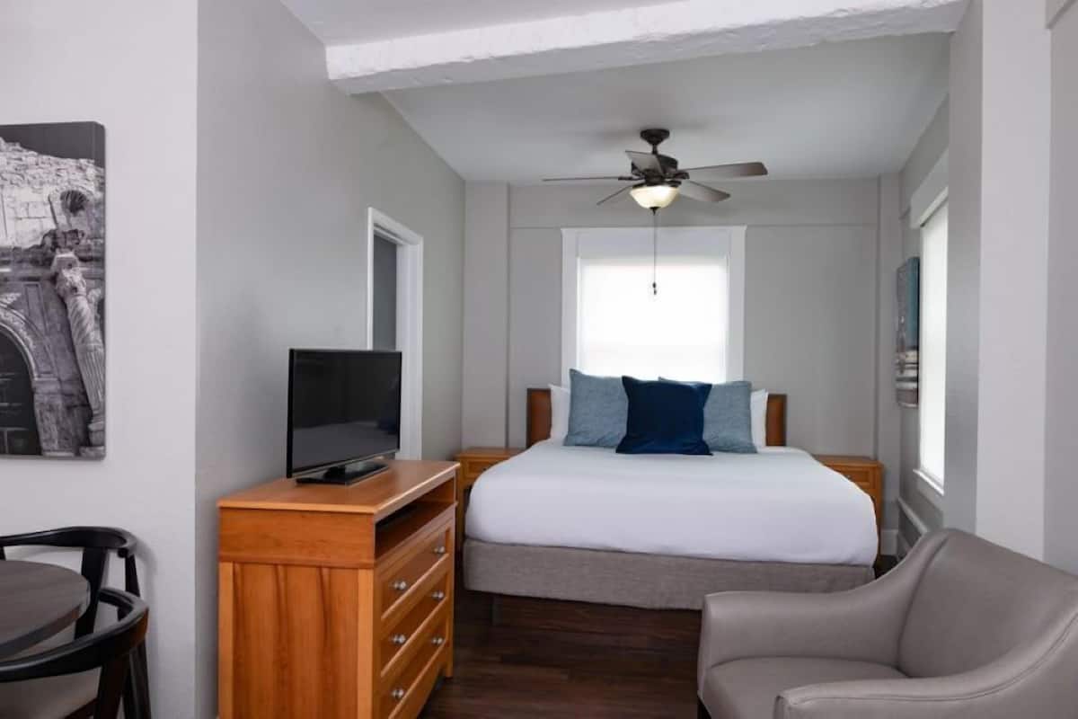 A bedroom with a bed, a TV on a wooden dresser, a ceiling fan, and a window.