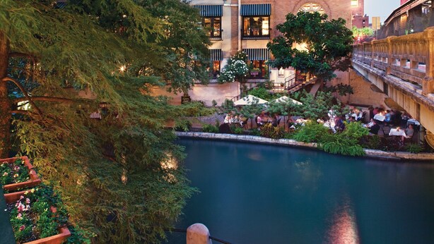 A riverside courtyard with outdoor seating, a brick building with multiple windows, and a bridge in the background. - Club Wyndham Riverside Suites (San Antonio)