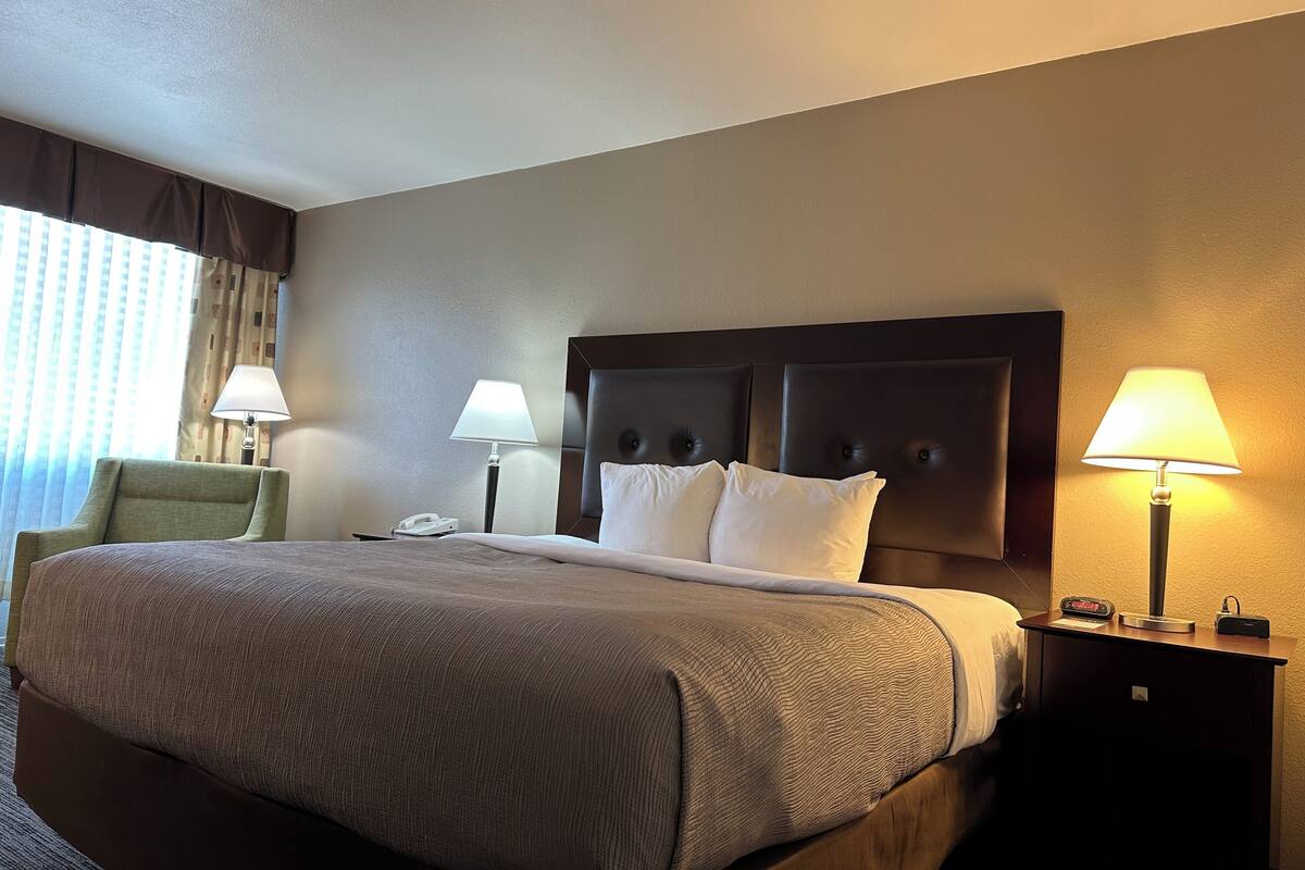 A hotel room with a large bed, two bedside lamps, a chair, and a nightstand with a phone and a clock.